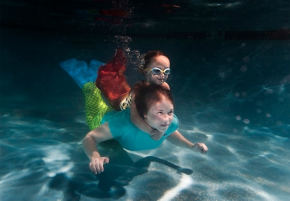 First Place, Portrait Personality - Logan Riely / Ohio UniversityCharlotte Echols, 5, holds onto the neck of her swim instructor Julia Mortenson as they cross the pool underwater in mermaid swimsuits at the Mermaid Swim School in Colorado Springs. The swim school offers mermaid classes to little kids and a monthly siren swim for adults who want to play in a pool wearing a mermaid tail.