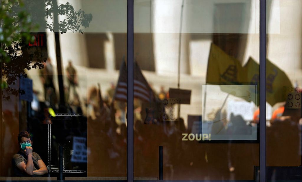 General News - 1st place - Protesters wave flags and signs in the reflection of the window at Zoup! on High Street outside of the Ohio Statehouse while Ohio Governor Mike DeWine and Ohio Department of Health Director Dr. Amy Acton give update on the status of the Coronavirus and the state's response on April 20. A crowd of over 300 protesters gathered outside the statehouse.   Kyle Robertson / The Columbus Dispatch