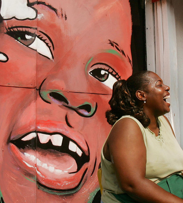 First Place, Photographer of the Year - John Kuntz / The Plain DealerLinda Taylor,42, owner of Lynn's Deli on Hough Avenue at the intersection of E. 82nd Street, laughs with business associate Gerald Mitchell outside the deli as they cook barbecued chicken for their lunch menu July 14, 2006.  The painted mural was done by a Nigerian artist who worked a portrait of her father King George Taylor who owned the property in the late 1960's and an unidentified girl laughing.  