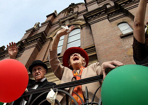 First Place, Photographer of the Year/Large Market - Lisa DeJong / The Plain DealerMarie Calandra, 88, and her husband retired Judge Salvatore Calandra, 88, on left, wave to the passing parade as they stand on the steps of the Holy Rosary Catholic Church during the Columbus Day Parade in Little Italy. The Calandras are the Grand Marshals of this year's parade. 