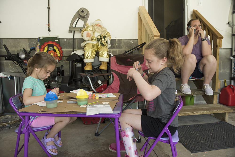 Second Place, Larry Fullerton Photojournalism Scholarship - Alexandria Skowronski / Ohio UniversityMelissa Smith (right) works over the phone to manage the Ultimate Wash car washes in Northeast Ohio while her children Maddie (left) and Sophia (center) play during their garage sale on May 18, 2019. Melissa works from home when she can because she takes Sophia to her physical therapy appointments, as many as 3 times a week. 
