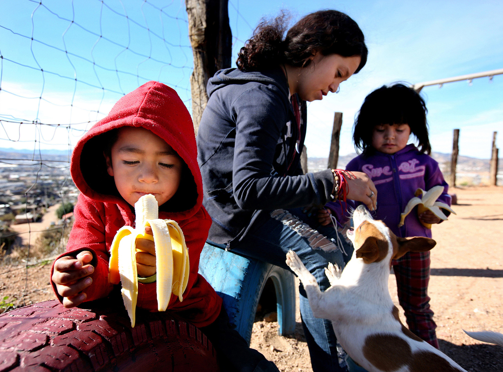 Second Place, News Picture Story - Lisa DeJong / The Plain Dealer, “Border Wall”Neythan Bermudez, 4, left, eats a banana, as his sister  Naydeiyn Bermudez, 15, and his cousin Ayiyn Bermudez, 3, play with a stray dog at Escuela Biblica N.A.N.A. in a neighborhood that lives next to a dump high above Nogales, Sonora, Mexico. The N.A.N.A. Ministry, a Hispanic title which translates to Children Helping Grateful Children, is a non-profit organization that feeds and houses children in the poor neighborhood of Rosarito Dos. Volunteers bring food daily. The need is great, as many of the children's parents have left to cross the border into the United States. 