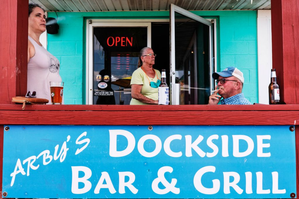 First Place, Chuck Scott Student Photographer of the Year - Carlin Stiehl / Ohio UniversityArby Holland smokes a cigarette on the patio with Sharlene Thornton, while his wife Debbie peaks outside to check on customers at Arby’s Dockside Grill on Deal Island, Md. Like many fishing towns, Arby’s is the only location on Deal Island where locals can get food and supplies. Since the pandemic began, shelves of tackle remain empty, but the store also functions as a bar and grill that has been doing well because it is the “only place to sell” crabs, Holland said.