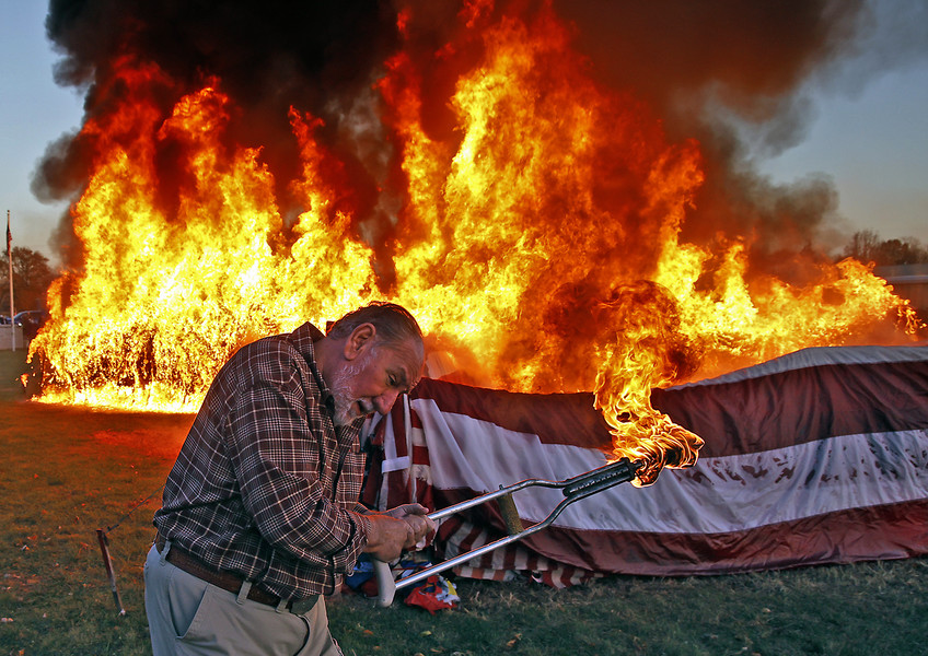 General News - 1st, “Flag Retirement”Lance James, a member of the American Legion Post 286 in New Carlisle, turns his face away from the heat as he uses a crutch to light 13,000 flags on fire during a Veterans Day Flag Retirement Ceremony Saturday, Nov. 11, 2023.  (Bill Lackey / Springfield News-Sun)
