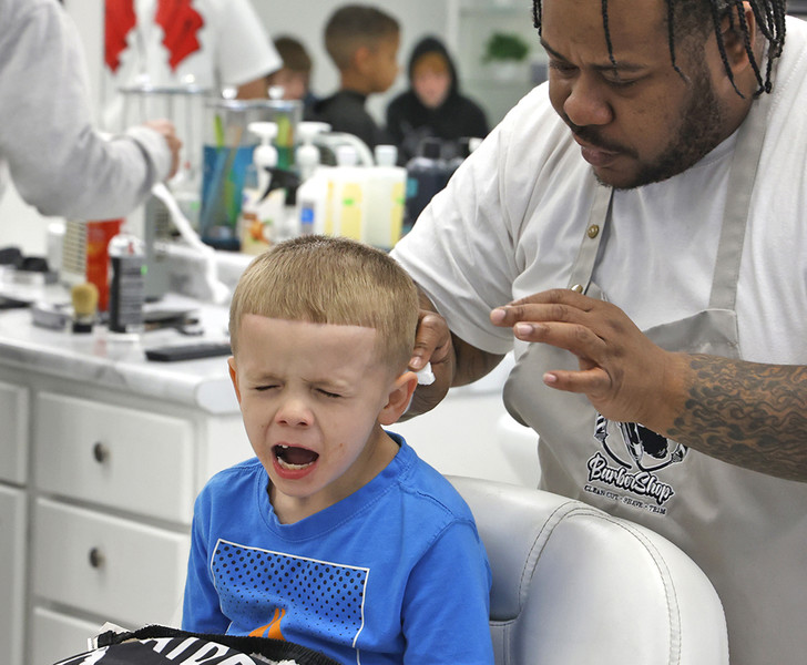 Award of Excellence - Photographer of the Year - Small Market Danny Riggins reacts as Dwain Garrett, one of the barbers at Champion City Cuts in the Southgate Shopping Center, brushes at hair off him after his haircut Monday, March 6, 2023.. The five barbers at Champion City Cuts volunteer their time the first Monday of every month to give Springfield City School students of all ages a fresh cut and some pizza as they wait their turn in the chair. The haircuts are offered as an incentive by the school district each month for good attendance.  (Bill Lackey / Springfield News-Sun)