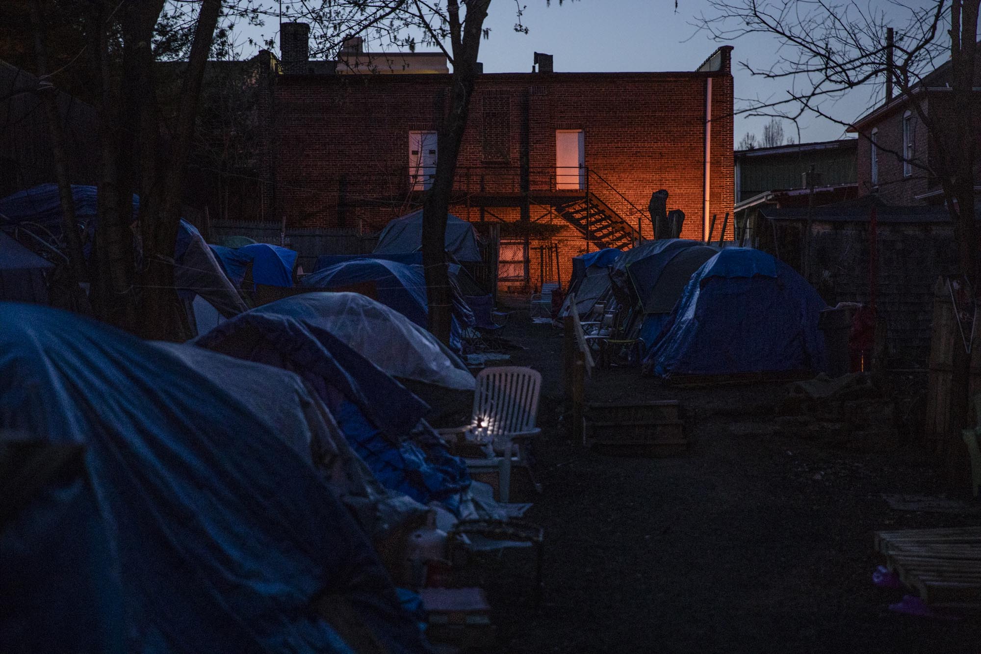 The sun sets on Tent City, showing light coming from security lamps installed by the low income apartment building next door. Multiple complaints about Tent City were filed by residents of that building, ranging from smells, to fights, to drug use and theft.